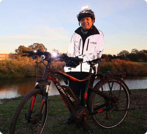 A rider stands with an e-bike by a river at dusk.  The rider is wearing reflective gear and a headlamp on his helmet.
