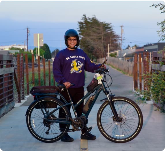 Smiling rider with a UC Santa Cruz sweatshirt stands behind e-bike.  In the background is a paved trail, one story buildings, and trees.
