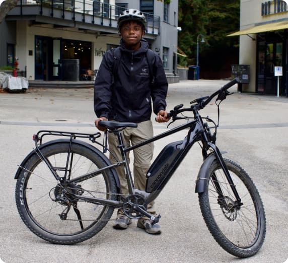 A rider wearing a light grey helmet and black jacket stands behind their e-bike in a spacious courtyard, with a two-story restaurant in the background.

